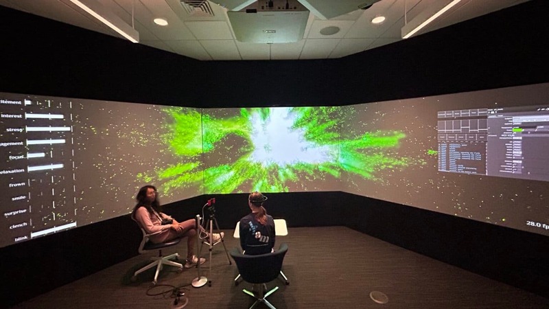Two people sit in the center of a dark room surrounded by walls of screens.