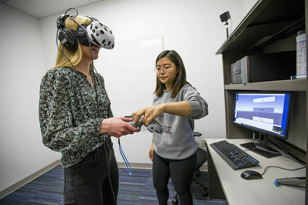A young woman has a black and white VR headset while a student worker is standing next to her helping her get adjusted to using the controllers in her hands.