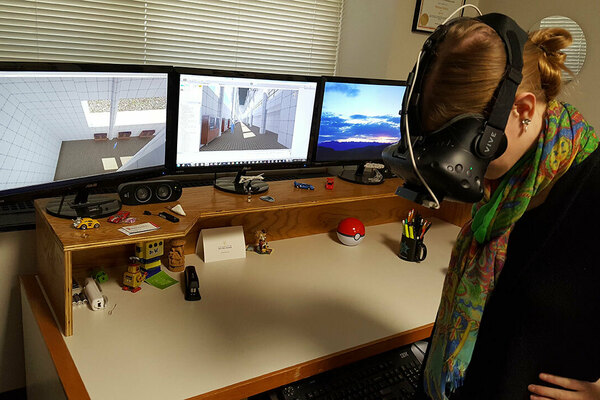 A young woman is looking down while wearing a set of black virtual reality google. In front of her are three computer monitors that display the content inside the VR goggles.